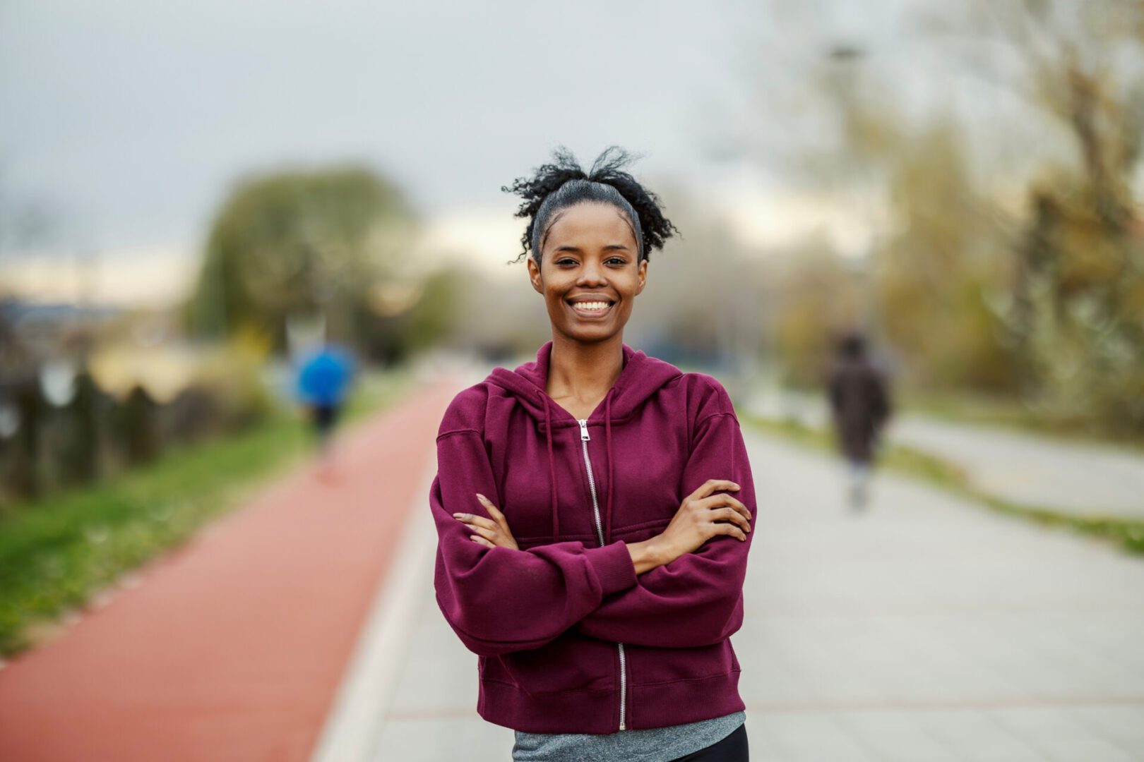 Young adult woman in a hoodie and sportswear standing with arms crossed, smiling at the camera on an outdoor running track
