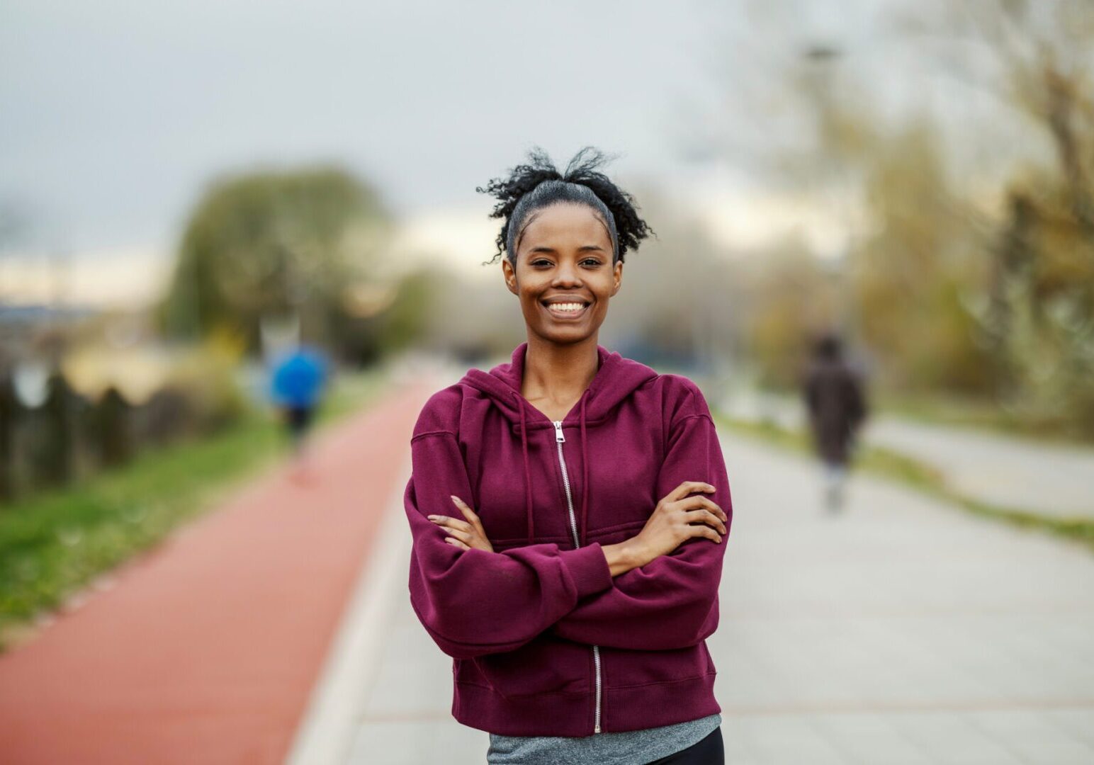 Young adult woman in a hoodie and sportswear standing with arms crossed, smiling at the camera on an outdoor running track